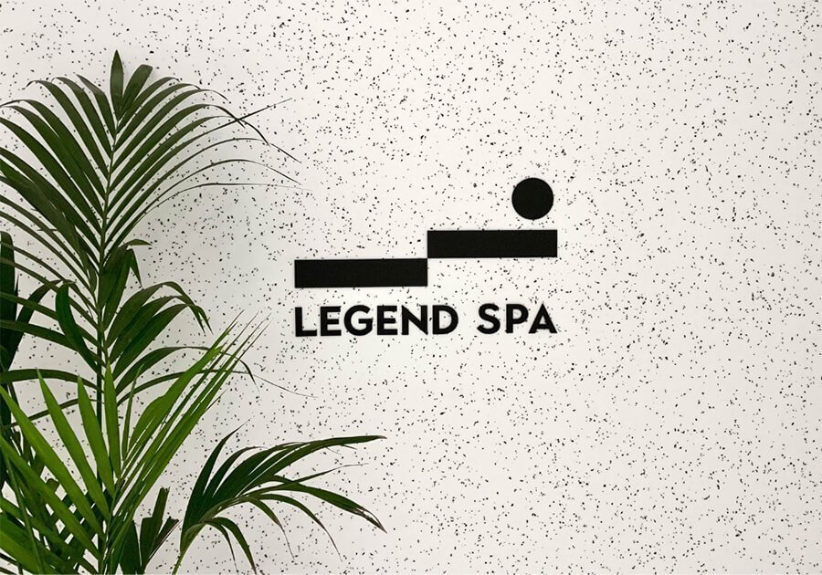 LEGEND Spa logo on speckled white wall with green plant beside it.