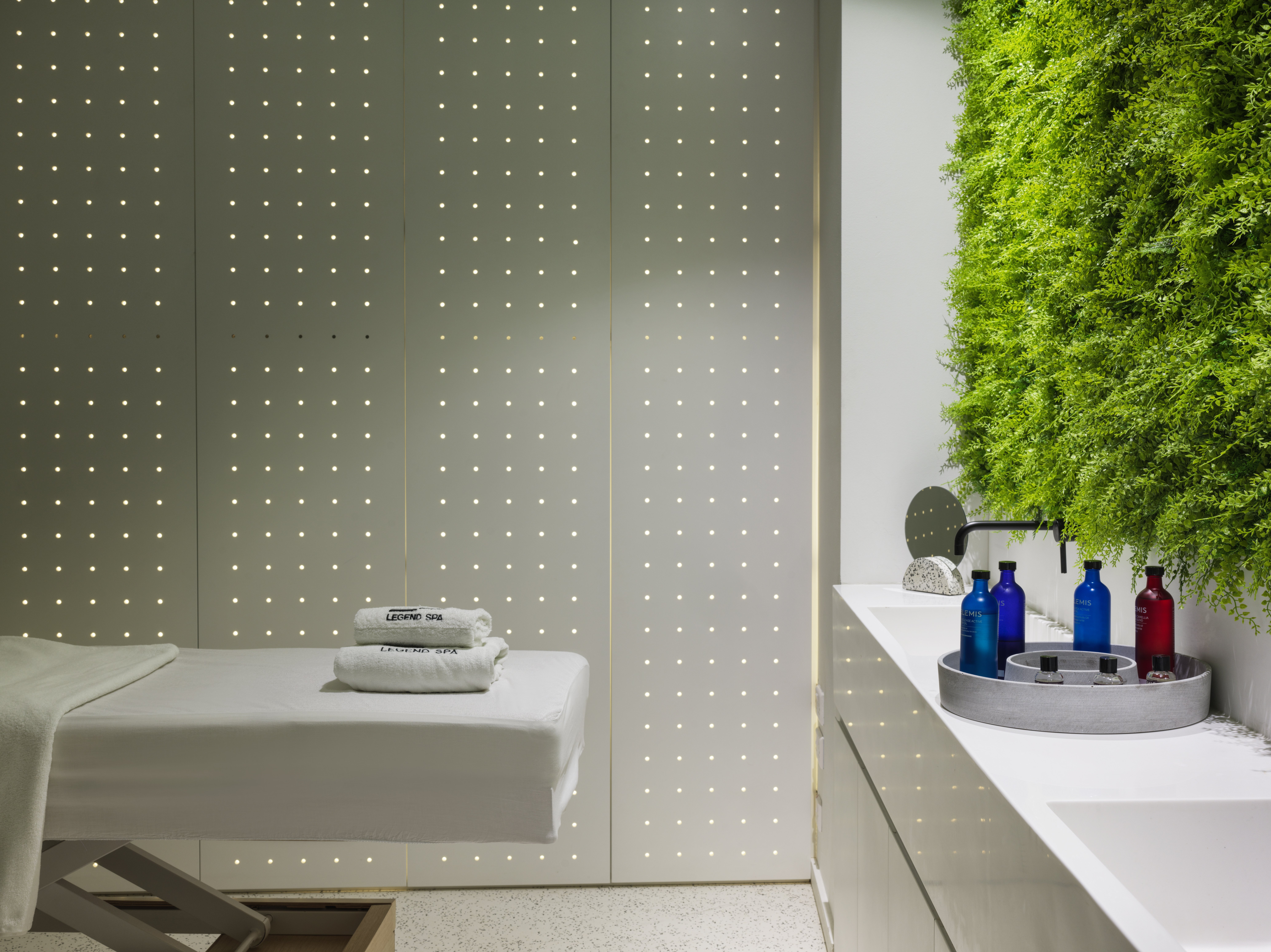 Modern spa room with massage table, soft lighting and plant wall.