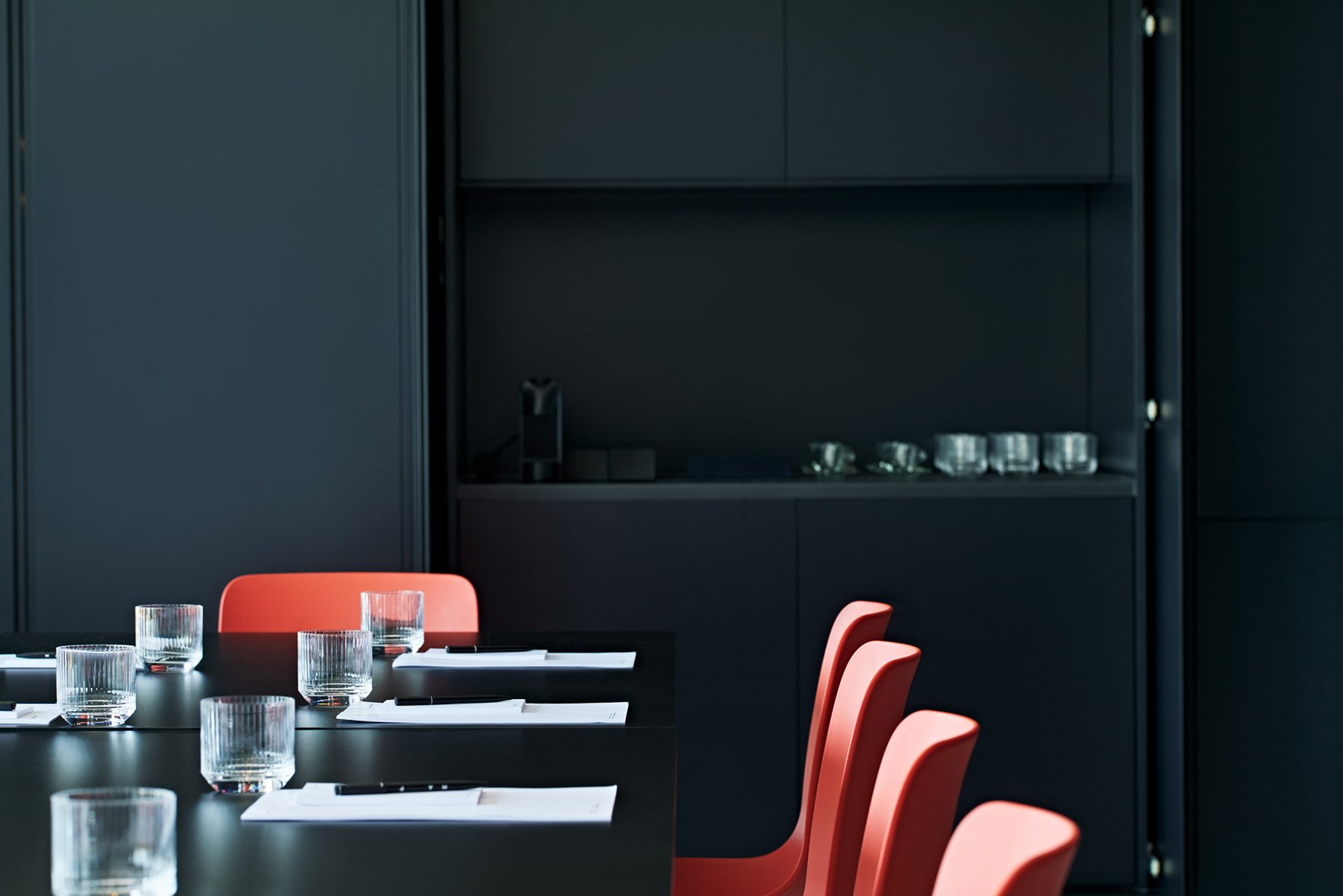 Meeting table set with red chairs, water glasses and black wall behind.