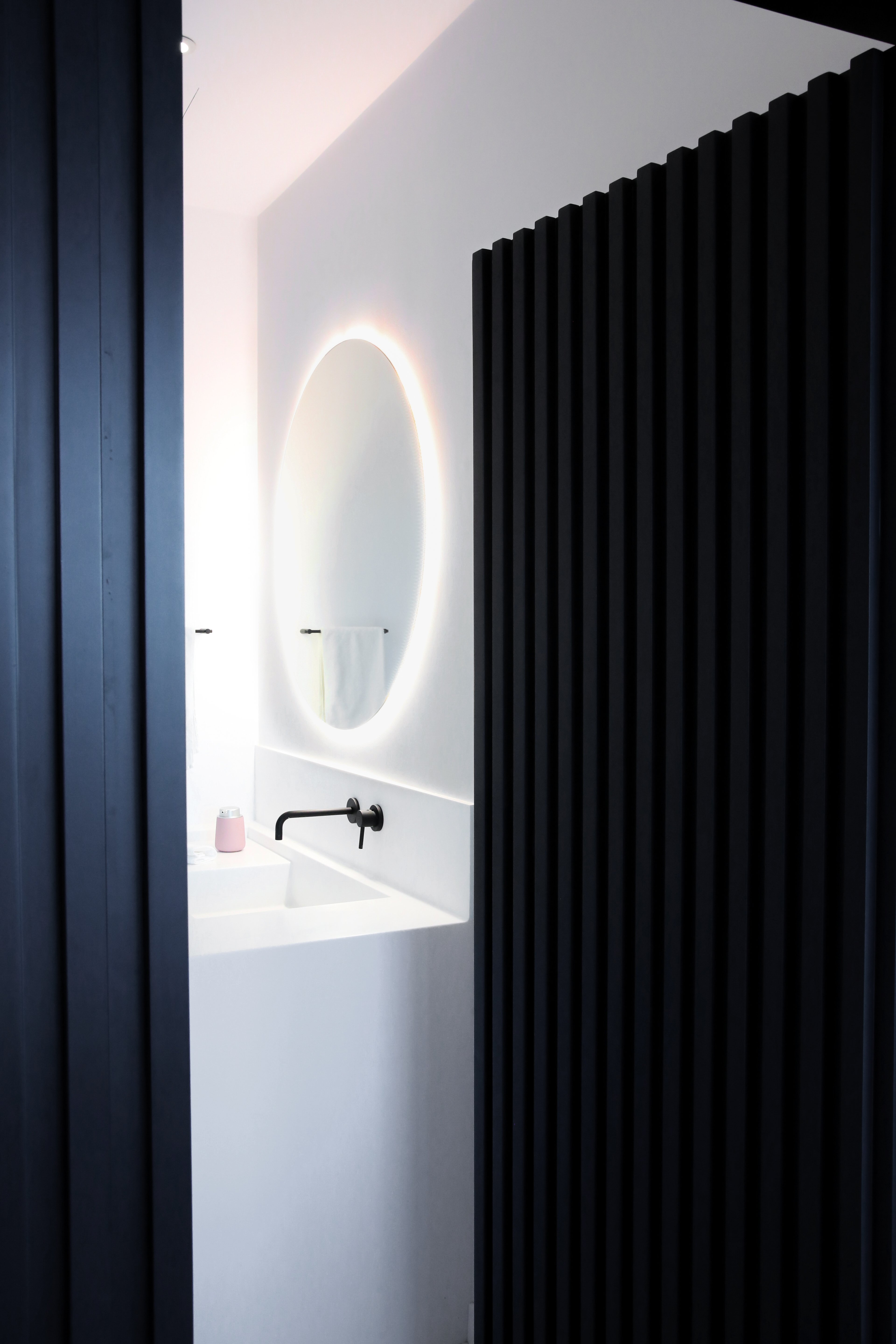Bathroom with round mirror and soft lighting behind dark slatted wall.