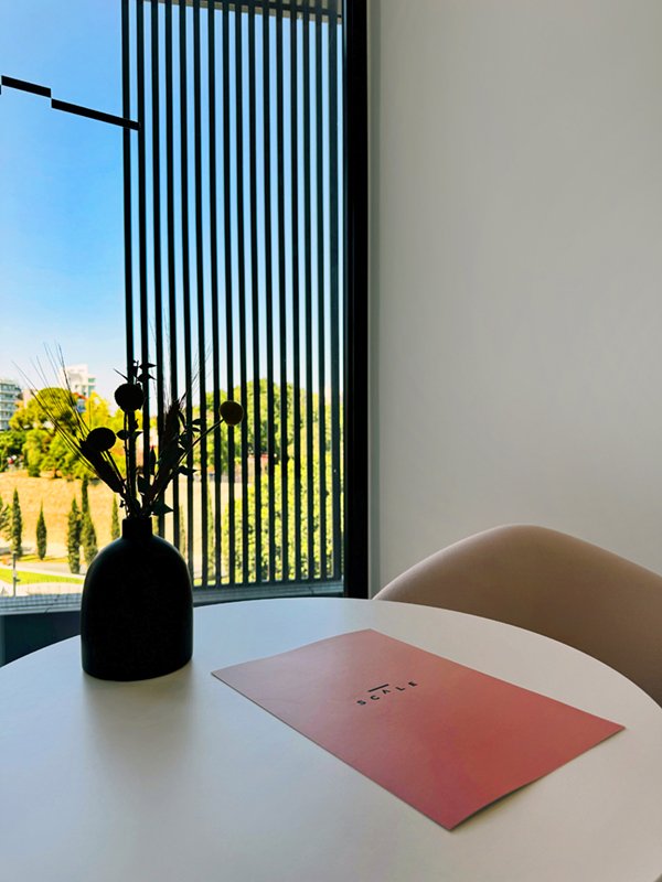 Small round table with pink folder and vase, sunlight through vertical blinds.