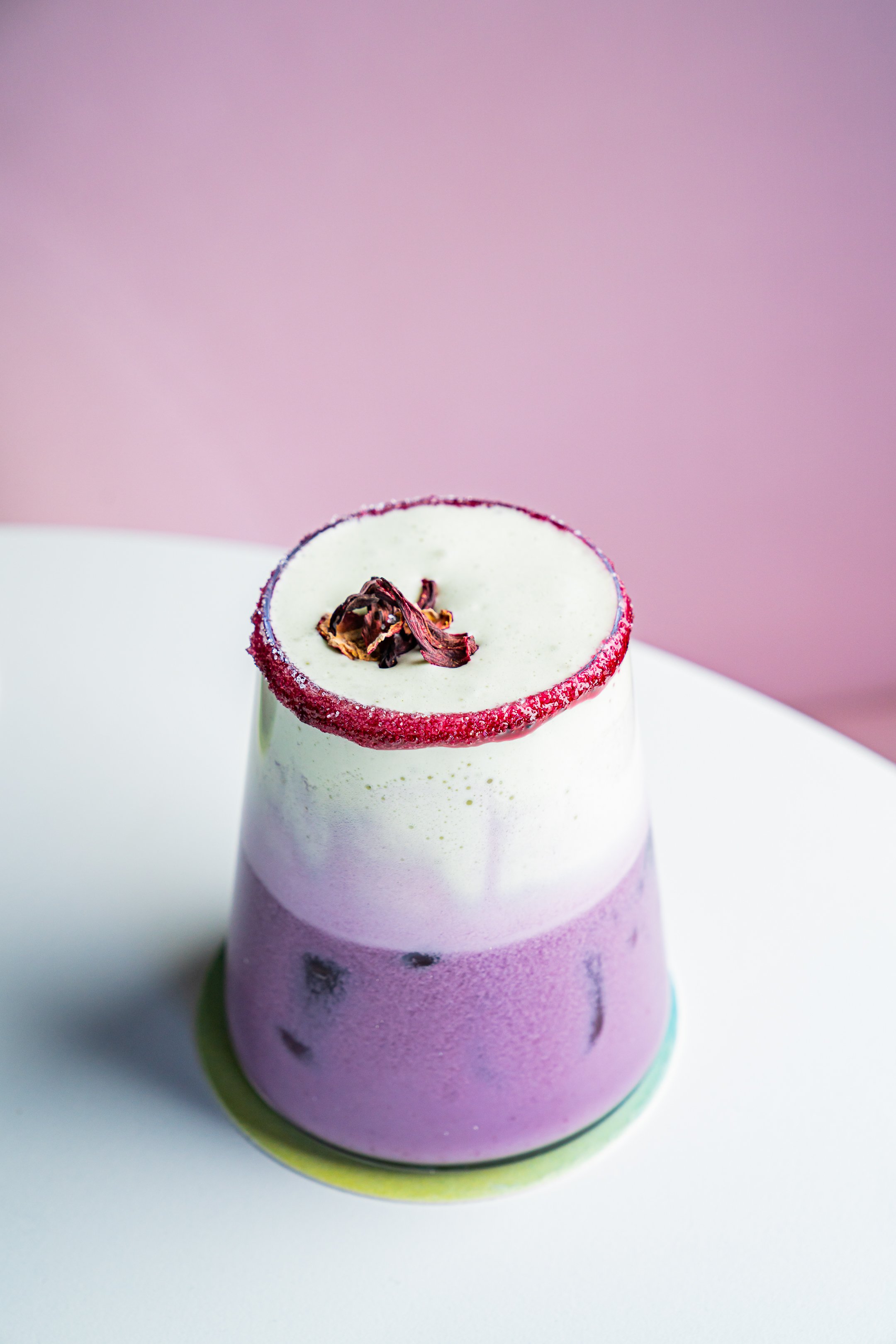 Violet smoothie with cream layer and flower garnish on pink table.