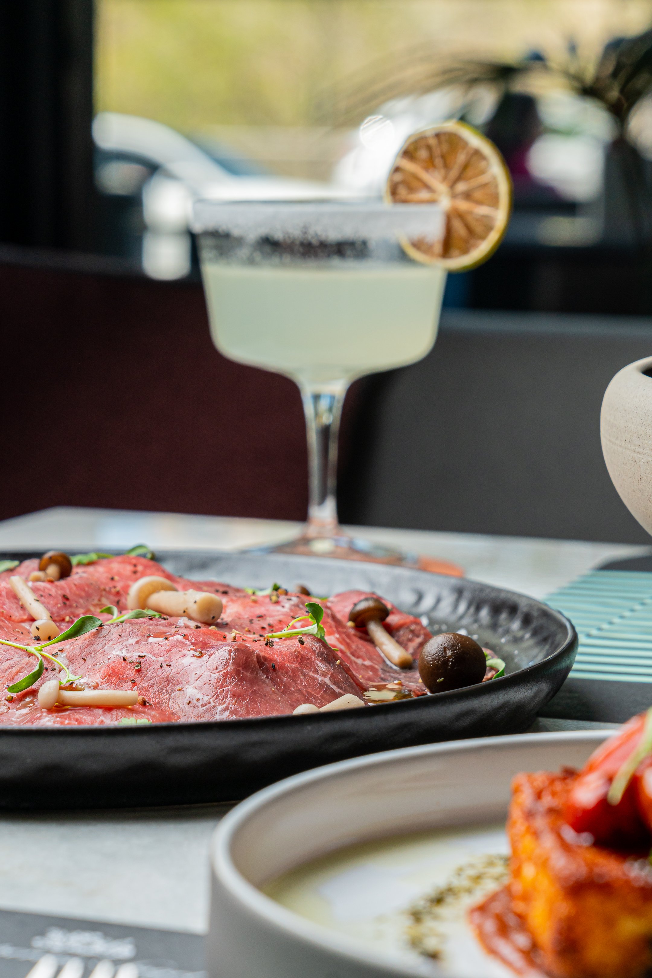Cocktail with lime slice beside plate of carpaccio.