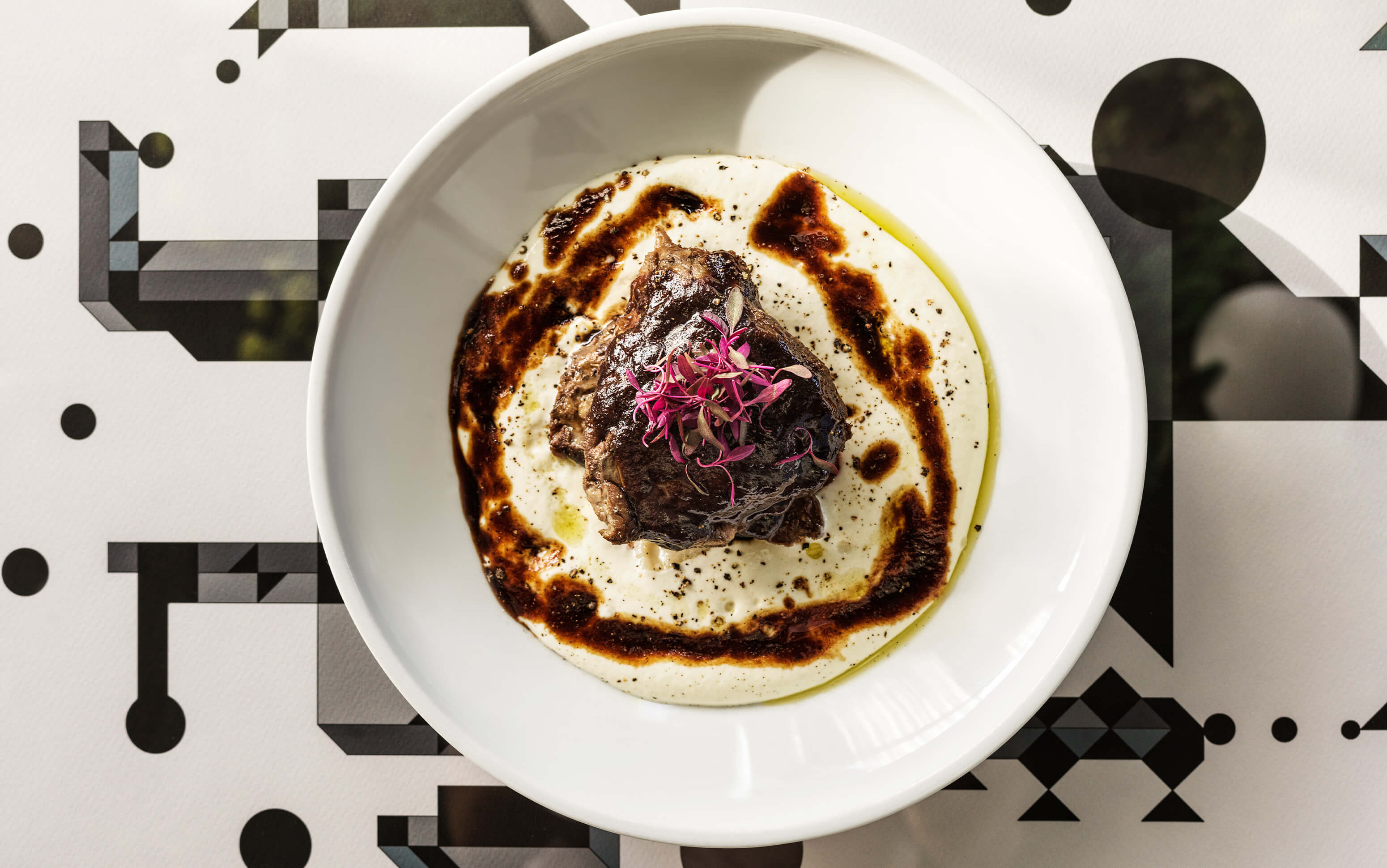 Gourmet dish with creamy puree, herbs and dark sauce served on patterned table.