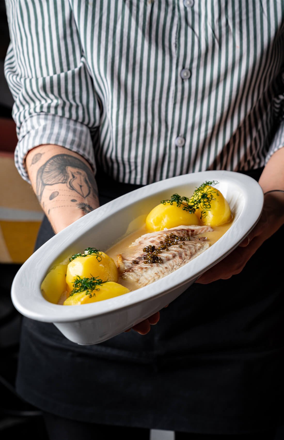 Chef presenting dish of fish fillet with yellow potatoes and herbs.