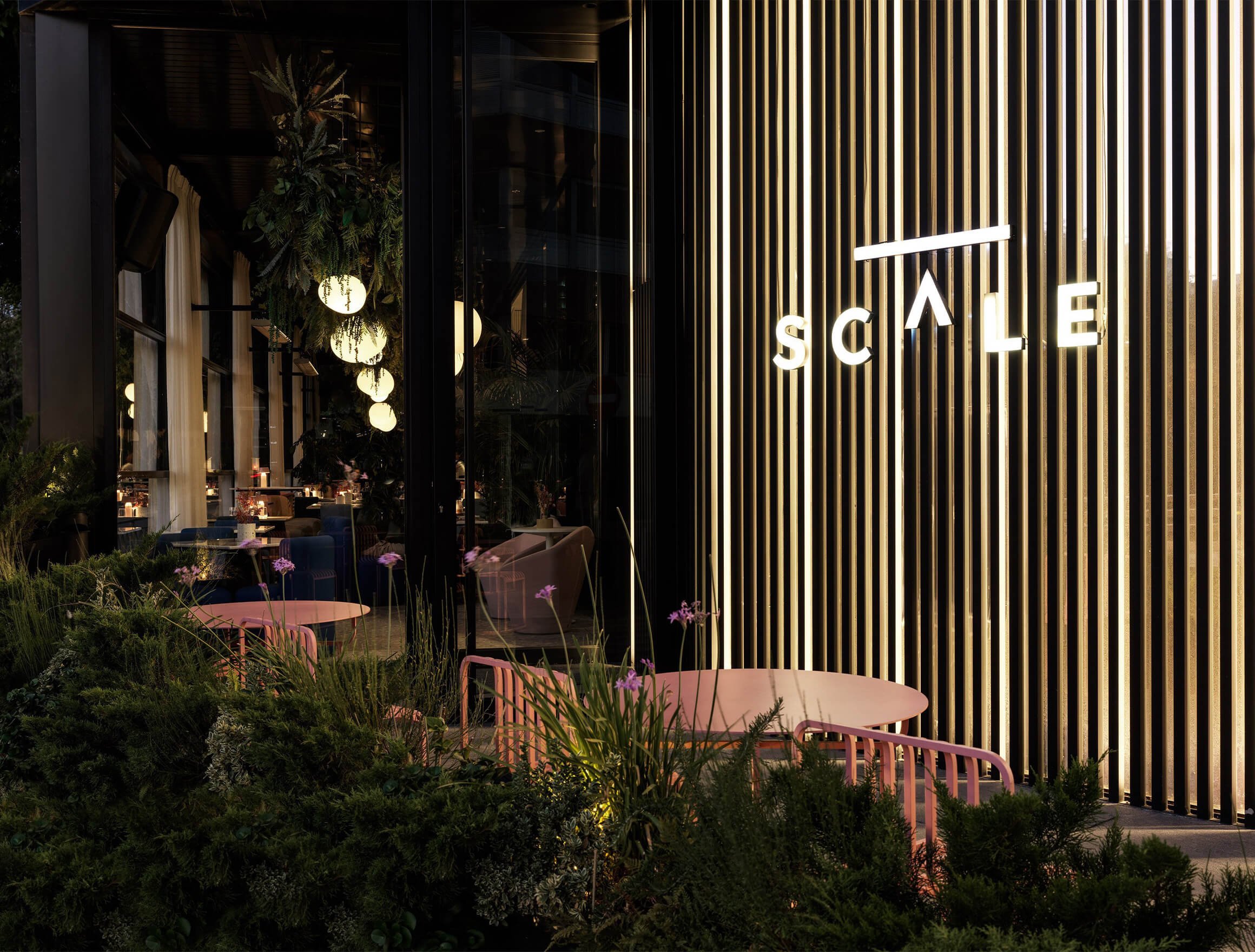 Exterior of SCALE restaurant entrance surrounded by plants and evening light.